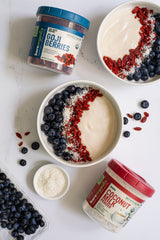 Bowl of yogurt with goji berries, blueberries, and coconut powder on a white surface.