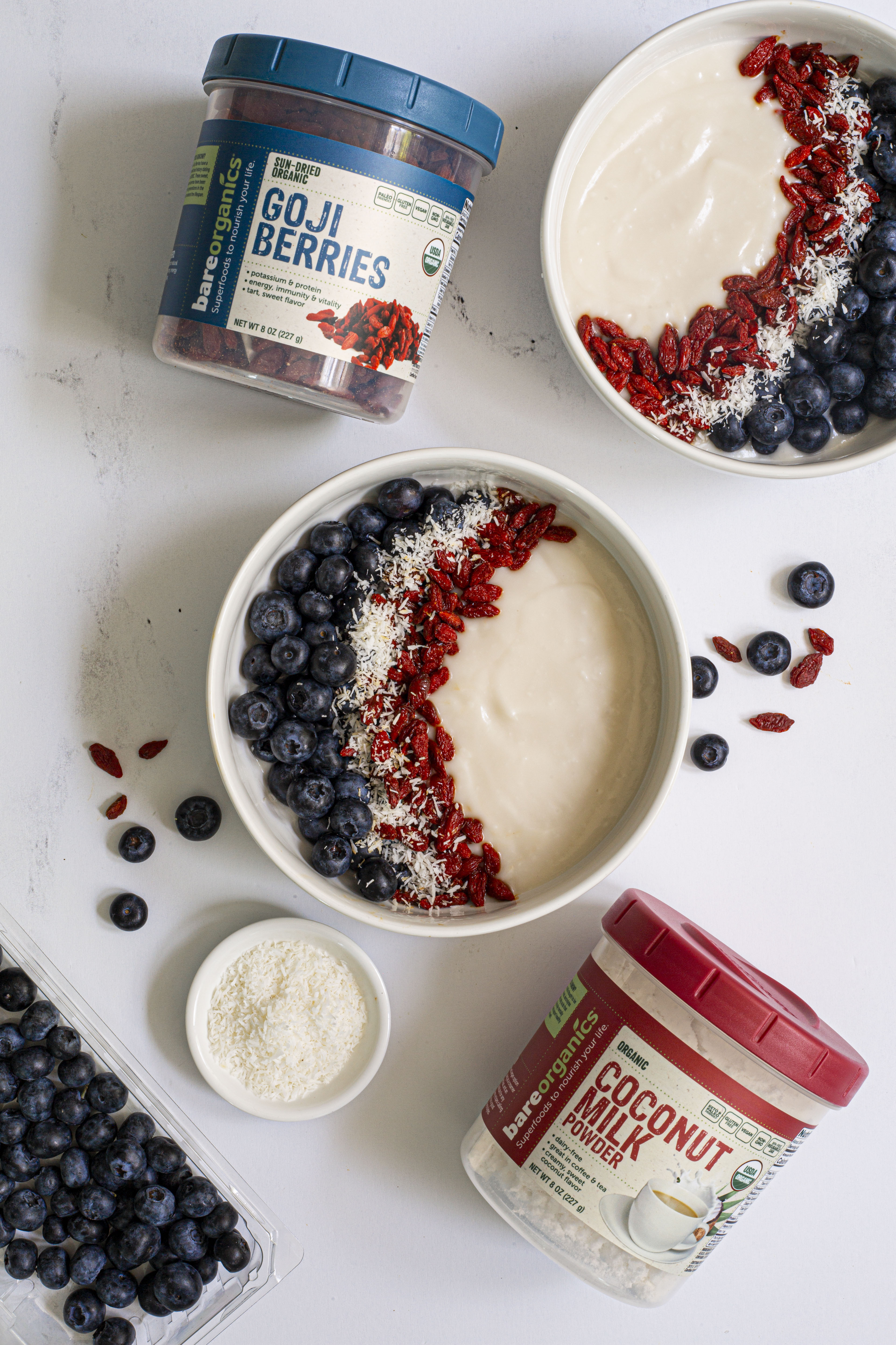 Bowl of yogurt with goji berries, blueberries, and coconut powder on a white surface.